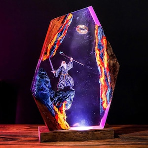May include: A decorative resin lamp featuring a wizard figure with a staff and sword, standing on a rocky base. The lamp has a wooden base and is encased in a clear resin with colorful accents of blue, orange, and yellow. A ring is suspended in the resin.