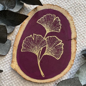 May include: A wooden round slice with a burgundy background and a gold ginkgo leaf design.
