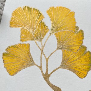 Könnte beinhalten: Aquarellmalerei eines Ginkgoblattzweigs. Die Blätter sind gelb mit goldenen Metallic-Akzenten, die Stiele sind braun. Das Kunstwerk befindet sich auf weißem Hintergrund.