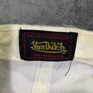 K&ouml;nnte beinhalten: Wei&szlig;e Baseballkappe mit einem schwarzen Etikett mit dem Text "Kustommade Von Dutch Originals TM" in goldfarbenen Buchstaben. Die Kappe hat ein klassisches Design und ist aus weichem Material gefertigt.