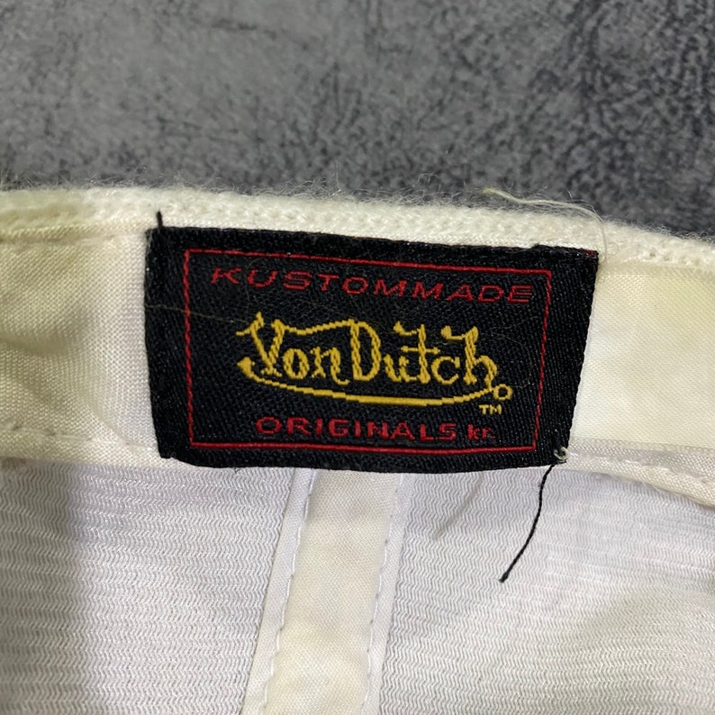K&ouml;nnte beinhalten: Wei&szlig;e Baseballkappe mit einem schwarzen Etikett mit dem Text "Kustommade Von Dutch Originals TM" in goldfarbenen Buchstaben. Die Kappe hat ein klassisches Design und ist aus weichem Material gefertigt.