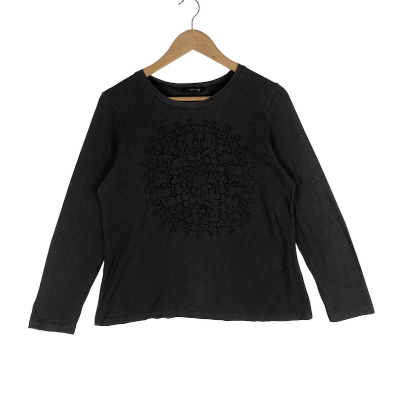 May include: Dark gray long-sleeve shirt with a crew neck. The shirt features a black floral mandala design on the front. The shirt is hanging on a wooden hanger against a white background.
