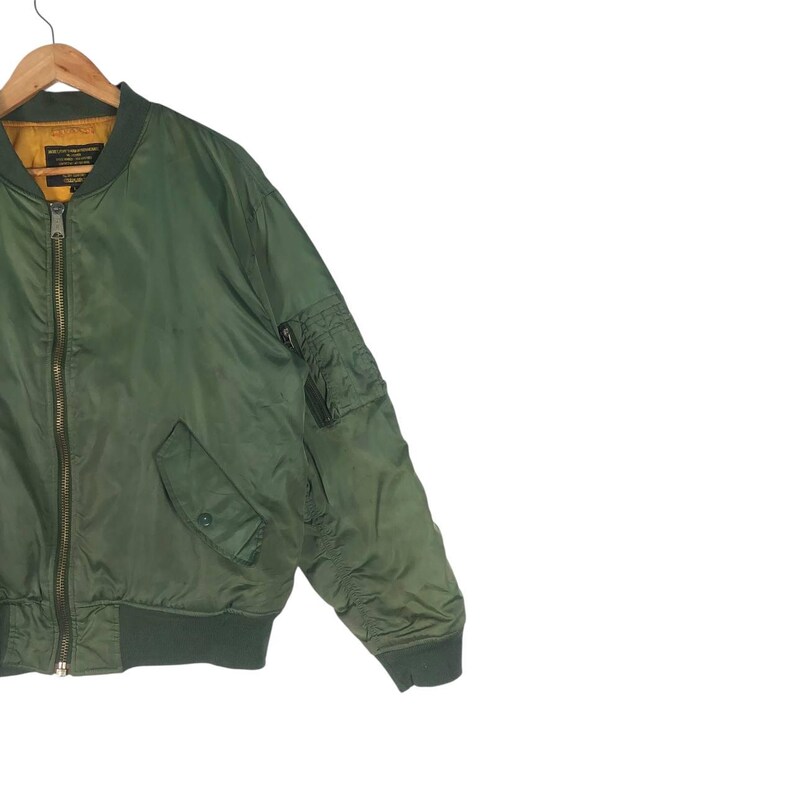 May include: Olive green bomber jacket with a ribbed collar, cuffs, and hem. The jacket features a zipper closure, two front pockets, and a utility pocket on the left sleeve. The jacket is hanging on a wooden hanger.