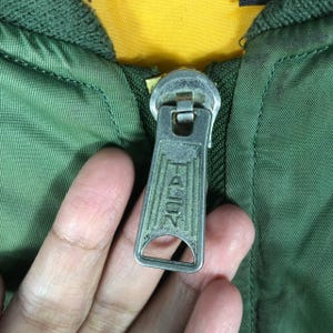 May include: Close-up of a green jacket zipper with a silver-colored metal pull. The zipper pull is embossed with the word "TALON". The jacket has a yellow lining.