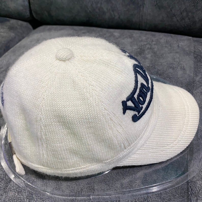 K&ouml;nnte beinhalten: Cremefarbene Baseballkappe mit gebogenem Schirm. Auf der Kappe ist das Wort "York" in marineblauer Schrift gestickt. Die Kappe besteht aus einem strukturierten Strickmaterial und hat einen kleinen Knopf oben.
