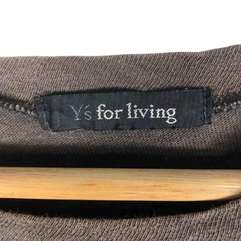 May include: Close-up of a brown garment with a black label. The label reads "Y's for living" in white text. The garment is hanging on a wooden hanger, and the fabric appears to be a knit material.