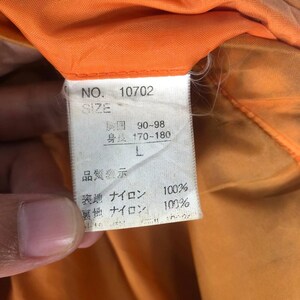 May include: Close-up of a tag with Japanese text and the number 10702. The tag indicates size L, with measurements of 90-98 for the chest and 170-180 for height. The tag is white with black text, set against an orange background.
