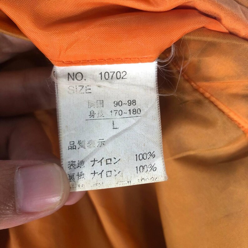 May include: Close-up of a tag with Japanese text and the number 10702. The tag indicates size L, with measurements of 90-98 for the chest and 170-180 for height. The tag is white with black text, set against an orange background.