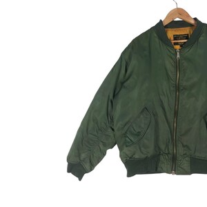 May include: A green bomber jacket with a full-length zipper, two front pockets, and ribbed cuffs and hem. The jacket is hanging on a wooden hanger against a white background. The jacket's color is a deep, muted green.