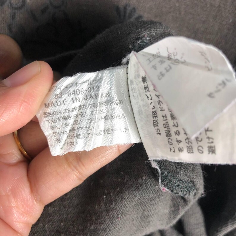 May include: Close-up of a gray garment tag with Japanese text and the words "MADE IN JAPAN". The tag is attached to a gray fabric. The text on the tag includes care instructions.
