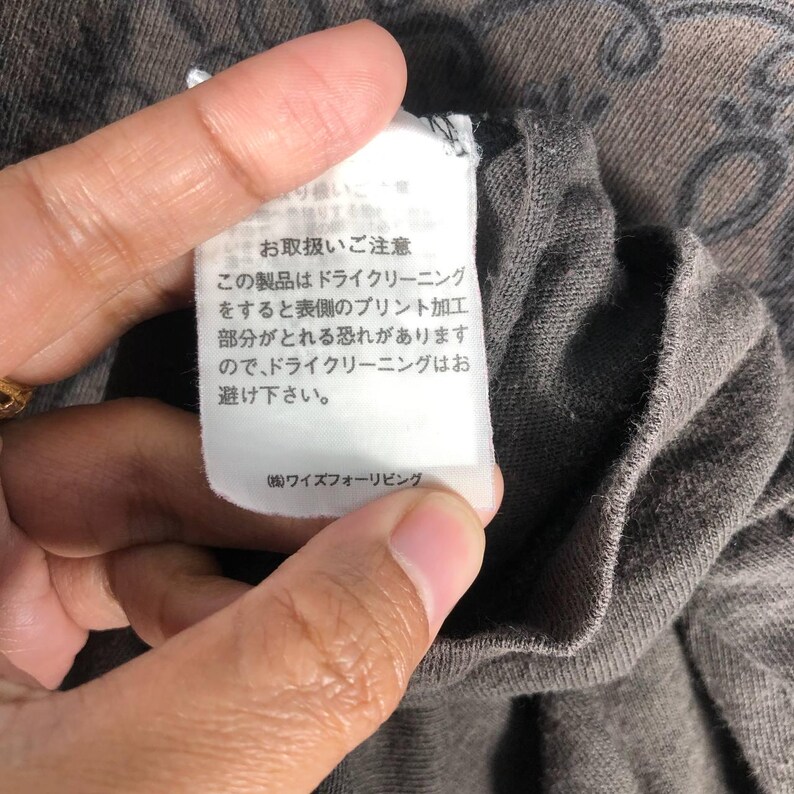 May include: Close-up of a gray t-shirt with a white tag. The tag has Japanese text, including care instructions about dry cleaning. The shirt's fabric is visible, along with a portion of a design.