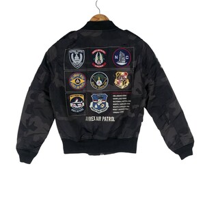 May include: Black camouflage bomber jacket with multiple patches featuring emblems and text, including "AVIREX AIR PATROL". The jacket has a ribbed collar, cuffs, and hem. Patches display state and regional names.