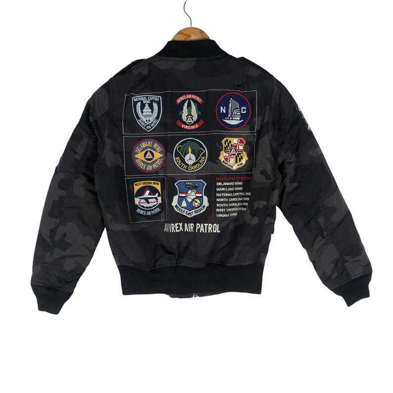May include: Black camouflage bomber jacket with multiple patches featuring emblems and text, including "AVIREX AIR PATROL". The jacket has a ribbed collar, cuffs, and hem. Patches display state and regional names.