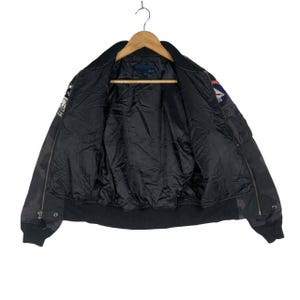 May include: Black bomber jacket with a camouflage design. The jacket features a black lining, a zip closure, and patches on the sleeves. It is suspended from a wooden hanger.