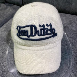 K&ouml;nnte beinhalten: Eine wei&szlig;e Baseballkappe aus Strick mit gebogenem Schirm. Auf der Kappe ist der Text "Von Dutch" in Marineblau aufgestickt. Die Kappe besteht aus einem strukturierten Strickmaterial.