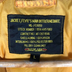 May include: Close-up of a yellow jacket label with black text. The label reads "JACKET, FLYR'S MAN INTRERMEDIATE" and includes details like the stock number, contact information, and material composition. The size is L.