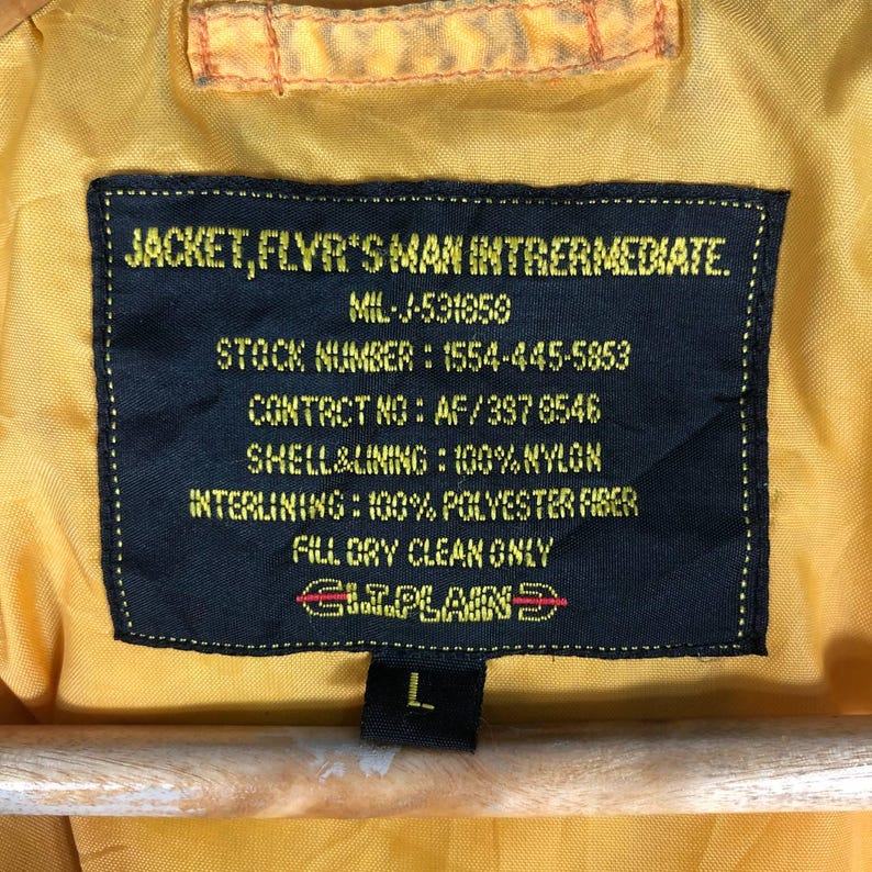 May include: Close-up of a yellow jacket label with black text. The label reads "JACKET, FLYR'S MAN INTRERMEDIATE" and includes details like the stock number, contact information, and material composition. The size is L.