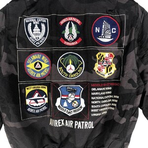 May include: Camouflage jacket in black with various embroidered patches. Patches feature text such as "National Capital Wing", "Avirex Air Patrol Virginia", "Delaware Wing", "South Carolina", "Maryland Wing", "West Virginia Wing", and "Middle East Region". The words "Avirex Air Patrol" are also present.