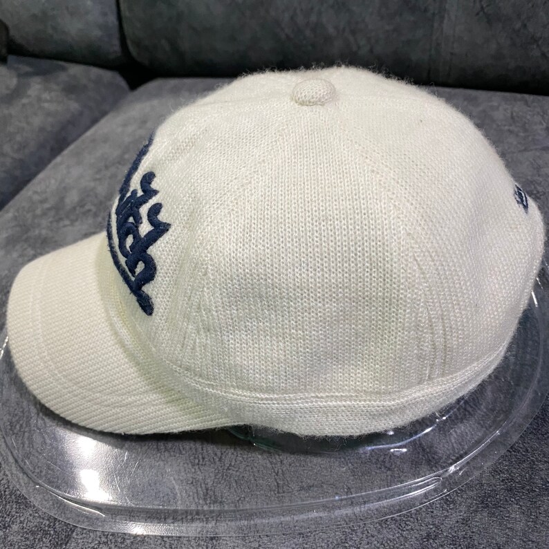 K&ouml;nnte beinhalten: Cremefarbene Baseballkappe mit gebogenem Schirm. Die Kappe hat ein marineblaues, gesticktes Design auf der Vorderseite und ein kleines Logo an der Seite. Sie besteht aus einem strukturierten Strickmaterial.