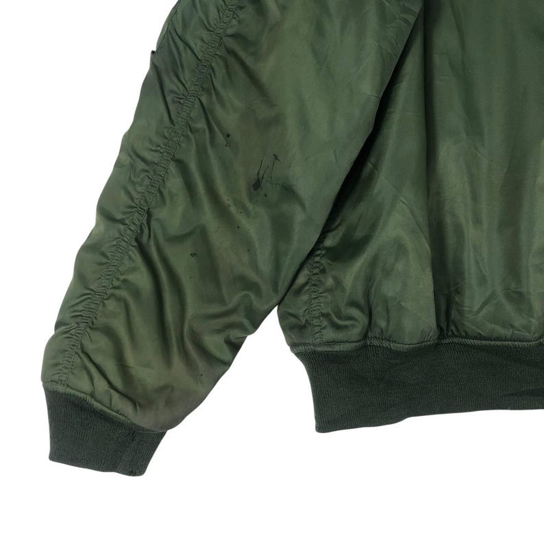 May include: Close-up of an olive green bomber jacket sleeve. The sleeve has a ribbed cuff and a quilted design on the upper arm. There are dark spots on the fabric. The jacket is a classic style.