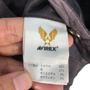 May include: A close-up of an Avirex garment tag. The white tag features the Avirex logo in gold and the brand name in black. The tag details the fabric composition: 62% acrylic, 27% wool, 9% polyester, and 2% polyurethane.