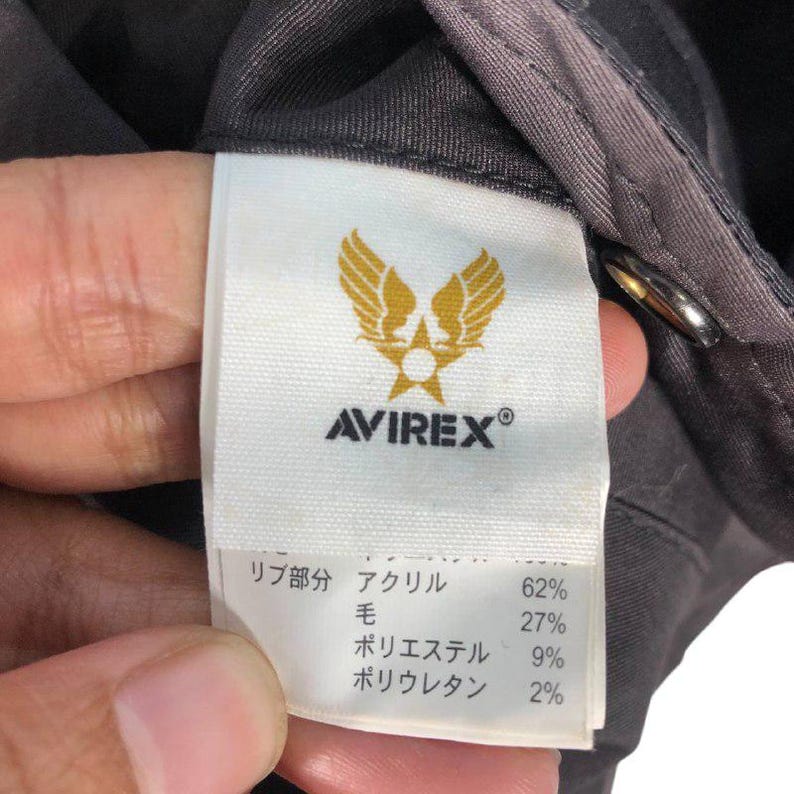 May include: A close-up of an Avirex garment tag. The white tag features the Avirex logo in gold and the brand name in black. The tag details the fabric composition: 62% acrylic, 27% wool, 9% polyester, and 2% polyurethane.