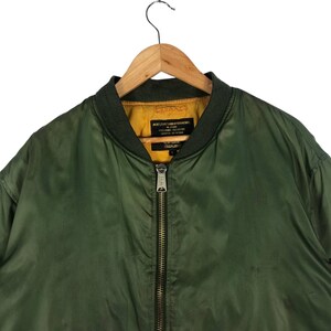 May include: Olive green bomber jacket with a dark green collar and a yellow interior. The jacket has a full-length zipper and a tag with text. The jacket is hanging on a wooden hanger.