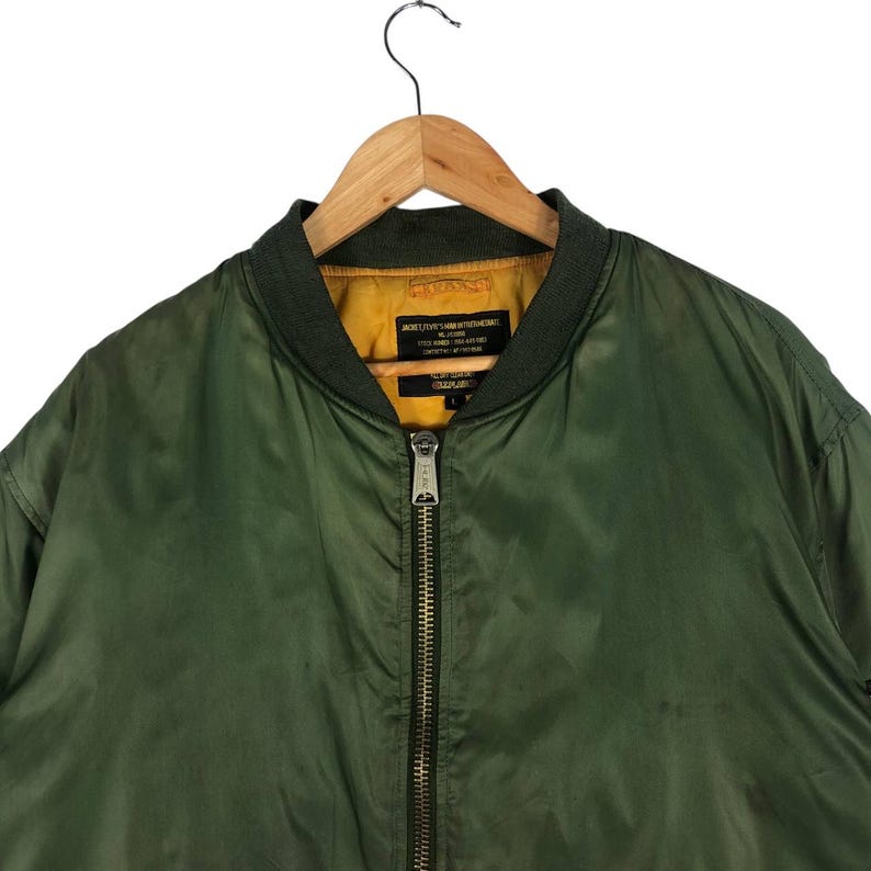 May include: Olive green bomber jacket with a dark green collar and a yellow interior. The jacket has a full-length zipper and a tag with text. The jacket is hanging on a wooden hanger.