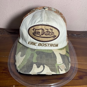 May include: A Von Dutch baseball cap with a white front panel and a brown oval logo. The brim is camouflage, and the mesh back is tan and brown. The text "ERIC BOSTROM" is embroidered below the logo.