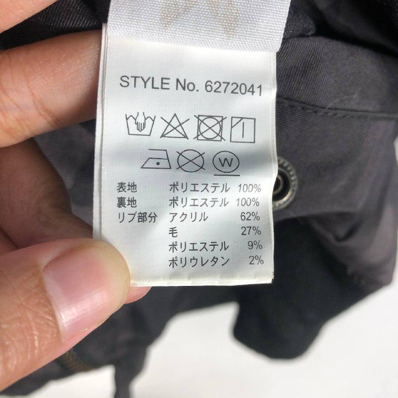 May include: A close-up shot of a white clothing label with the text "STYLE No. 6272041." The label details fabric percentages in Japanese. The tag is attached to a dark garment. The image is well-lit and in focus.
