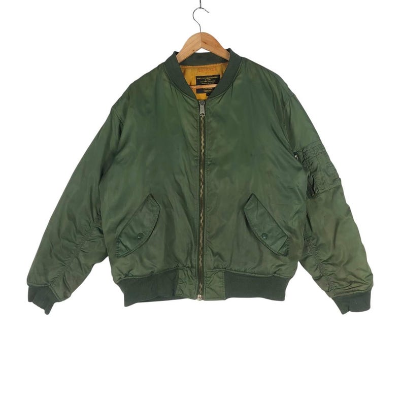 May include: Olive green bomber jacket with a zip-up front, two front pockets, and a pocket on the left sleeve. The jacket has a ribbed collar, cuffs, and hem. The interior lining is a golden yellow color.