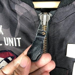 May include: Close-up of a black jacket zipper with a leather pull tab. The jacket features a camouflage pattern in shades of grey and white. The text "UNIT" is visible in white lettering.