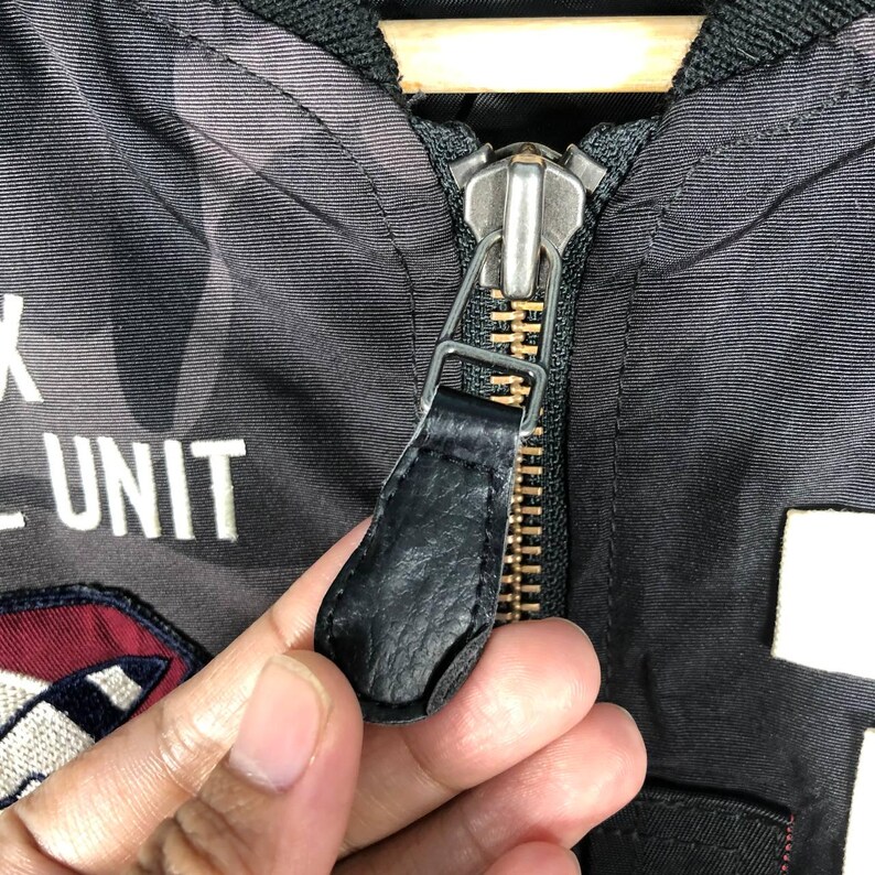 May include: Close-up of a black jacket zipper with a leather pull tab. The jacket features a camouflage pattern in shades of grey and white. The text "UNIT" is visible in white lettering.