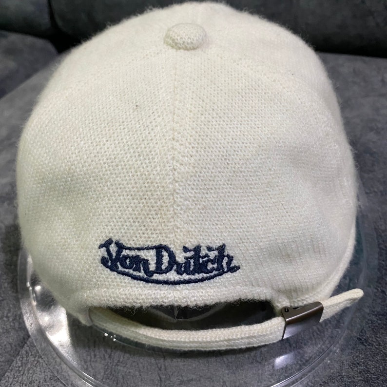 K&ouml;nnte beinhalten: Cremefarbene Strickm&uuml;tze mit gebogenem Schirm und kleinem Knopf oben. Der Schriftzug "Von Dutch" ist in Marineblau auf der R&uuml;ckseite gestickt. Die Kappe hat einen Metallverschluss zur Anpassung.