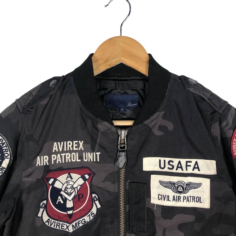 May include: A black camouflage bomber jacket with various patches. The jacket displays the text "AVIREX AIR PATROL UNIT" and "USAFA CIVIL AIR PATROL". It is suspended from a wooden hanger.