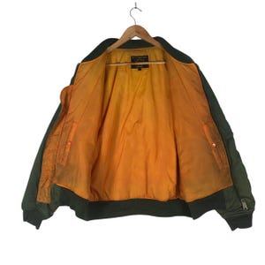 May include: Olive green bomber jacket with an orange lining, displayed open on a wooden hanger. The jacket has a ribbed collar, cuffs, and hem. Features a zipper closure and a small pocket on the left sleeve. A classic outerwear piece.