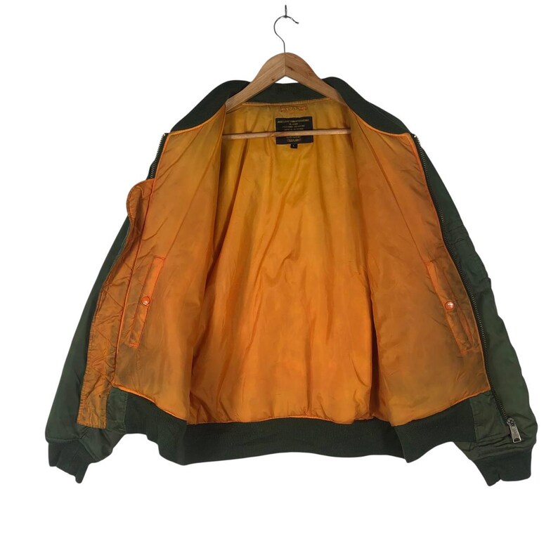May include: Olive green bomber jacket with an orange lining, displayed open on a wooden hanger. The jacket has a ribbed collar, cuffs, and hem. Features a zipper closure and a small pocket on the left sleeve. A classic outerwear piece.