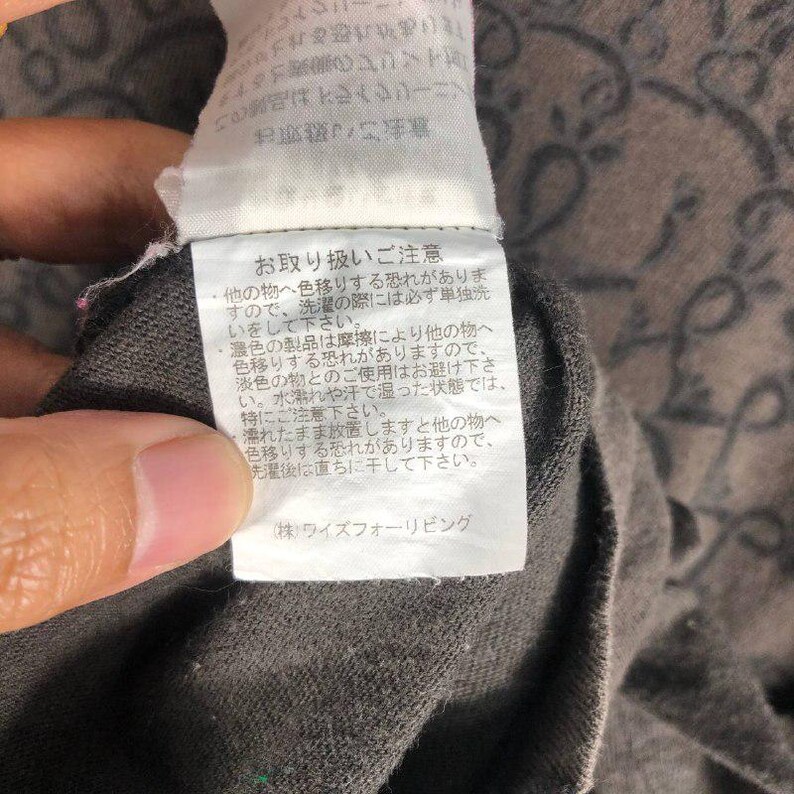 May include: Close-up of a gray fabric with a tag attached. The tag has Japanese text and is white with black text. The fabric has a subtle pattern. The image is well-lit and shows the texture of the fabric.