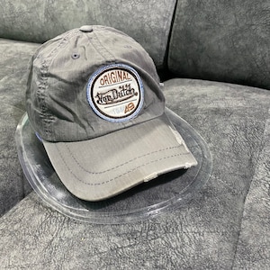 Puede incluir: Gorra de béisbol gris con visera desgastada. La gorra presenta un parche circular con el texto "ORIGINAL Von Dutch 1949" en blanco, marrón y azul.