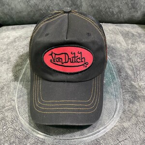 May include: Black baseball cap with a mesh back and a curved brim. The cap features the text "Von Dutch" in a red oval patch on the front. The stitching is gold-colored.