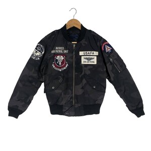 May include: Black camouflage bomber jacket with various patches, including "AVIREX AIR PATROL UNIT" and "USAF". The jacket has a black ribbed collar, cuffs, and waistband. Features two front pockets and a zip closure.