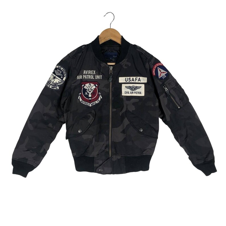 May include: Black camouflage bomber jacket with various patches, including "AVIREX AIR PATROL UNIT" and "USAF". The jacket has a black ribbed collar, cuffs, and waistband. Features two front pockets and a zip closure.