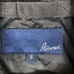 May include: Detail of a black jacket's lining, showing a navy blue label with the white cursive brand name "Avirex" and a small "S" size tag. The jacket's lining is a textured, dark fabric.