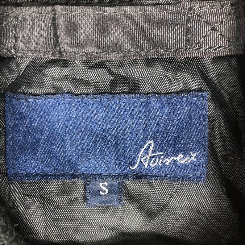 May include: Detail of a black jacket's lining, showing a navy blue label with the white cursive brand name "Avirex" and a small "S" size tag. The jacket's lining is a textured, dark fabric.