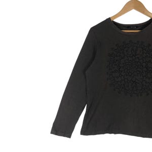 May include: A dark gray long-sleeve shirt with a black floral mandala design on the front. The shirt is hanging on a wooden hanger against a white background. The shirt has a crew neck.