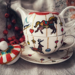 Może przedstawiać: Biały ceramiczny zestaw do herbaty z motywem konia z karuzeli. Zestaw zawiera spodek i pokrywkę. Czajniczek i filiżanka są ułożone w stos, a pokrywka jest umieszczona obok. Czerwone jagody w tle.