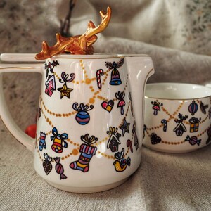 Könnte beinhalten: Ein weißes Keramik-Teeservice, verziert mit farbenfrohen Illustrationen zum Thema Weihnachten. Die Teekanne hat einen goldfarbenen Deckelgriff in Form eines Geweihs. Das Set ist mit festlichen Ornamenten, Zuckerstangen und Lebkuchenhäusern verziert.