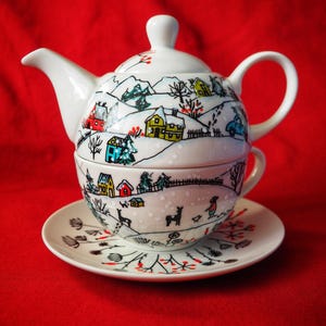 Może przedstawiać: Biały ceramiczny zestaw do herbaty z motywem zimowej wioski. Zestaw zawiera czajniczek, filiżankę i spodek. Czajniczek i filiżanka są składane. Projekt przedstawia kolorowe domy, drzewa i samochód na tle zaśnieżonego krajobrazu.