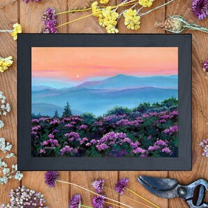 May include: A framed landscape painting depicting a field of purple flowers in the foreground with rolling blue mountains under a soft orange and pink sunset sky. The artist signature SKYMIR is visible in the lower left corner of the artwork.