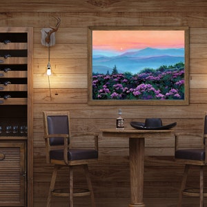 May include: A framed landscape painting depicting a sunset over blue mountain ridges and a foreground of vibrant purple rhododendron flowers. The artwork is displayed on a wood-paneled wall above a round wooden table with a bottle and a black hat.